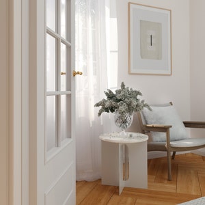 Op de afbeelding: Een witte bijzettafel met een wit marmeren blad en een vaas met witte bloemen erop. De tafel staat in een kamer met witte muren en een witte deur. Er ligt een wit tapijt op de vloer en een stoel met een wit kussen.