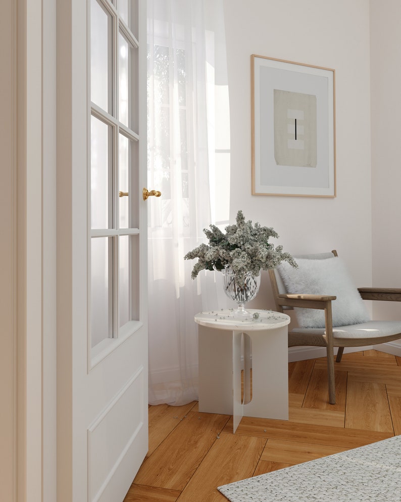 Op de afbeelding: Een witte bijzettafel met een wit marmeren blad en een vaas met witte bloemen erop. De tafel staat in een kamer met witte muren en een witte deur. Er ligt een wit tapijt op de vloer en een stoel met een wit kussen.