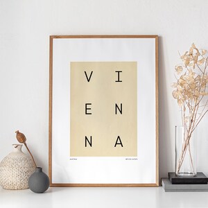 May include: A framed print with the word "VIENNA" spelled out in a grid pattern. The print is on a cream background with black text. The frame is a light brown wood.