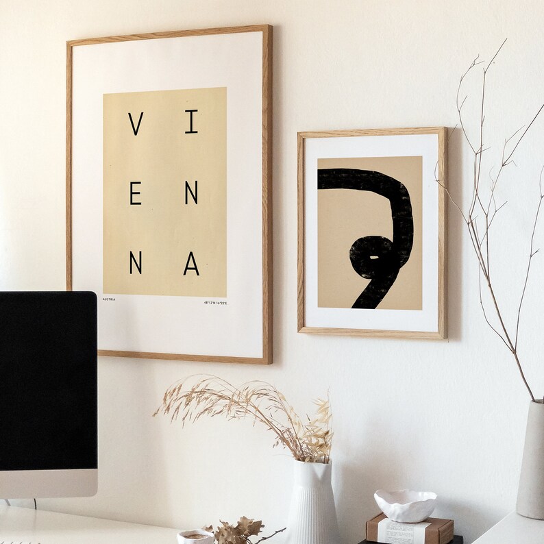 May include: Two framed prints on a white wall. The print on the left is a beige background with the word "VIENNA" in black letters. The print on the right is a beige background with a black abstract line drawing.