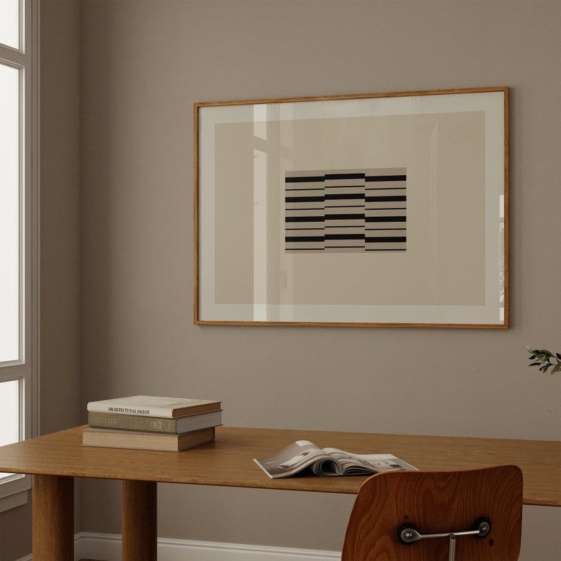 May include: A framed black and white geometric print with a beige background hangs on a light beige wall above a wooden desk with a stack of books and a magazine.