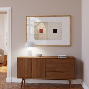 Op de afbeelding: Een houten dressoir met twee deuren en drie lades. Het dressoir heeft een lichte houtkleur en staat op een visgraatmotief houten vloer. Een witte lamp en een witte kom staan op het dressoir. Een ingelijst schilderij met een rood en zwart vierkant ontwerp hangt aan de muur achter het dressoir.