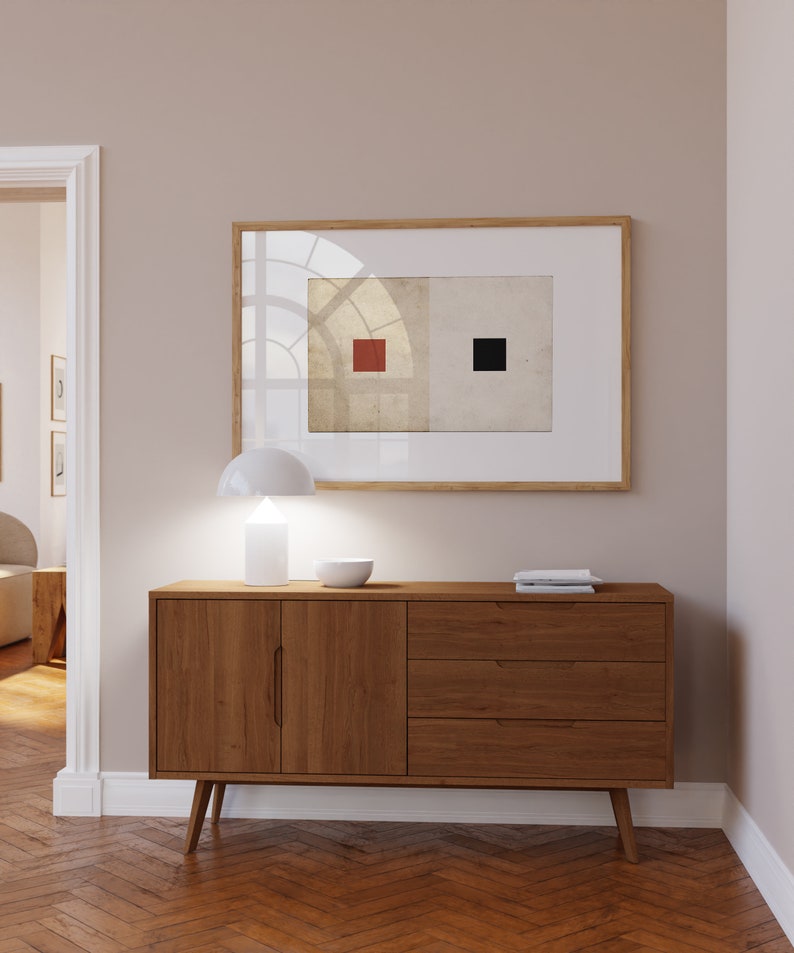 Op de afbeelding: Een houten dressoir met twee deuren en drie lades. Het dressoir heeft een lichte houtkleur en staat op een visgraatmotief houten vloer. Een witte lamp en een witte kom staan op het dressoir. Een ingelijst schilderij met een rood en zwart vierkant ontwerp hangt aan de muur achter het dressoir.