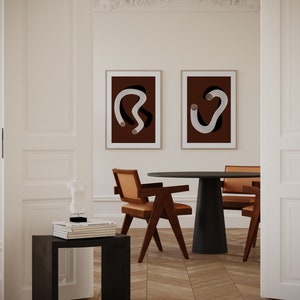 Könnte beinhalten: Zwei gerahmte abstrakte Kunstdrucke mit weißen Linien auf einem dunkelbraunen Hintergrund hängen an einer weißen Wand über einem schwarzen runden Tisch mit vier braunen Stühlen. Ein kleiner schwarzer Beistelltisch mit einer weißen Skulptur und Büchern steht in der Ecke des Raumes.