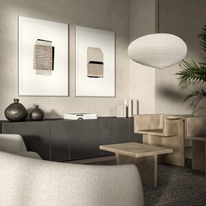 May include: Two framed abstract art prints with beige and black paper textures hang on a light beige wall above a dark gray cabinet. A white, round, layered pendant light hangs from the ceiling. A white, curved sofa sits in front of the cabinet. A light brown, rectangular coffee table sits in front of the sofa. A light brown, abstract sculpture sits on the floor.