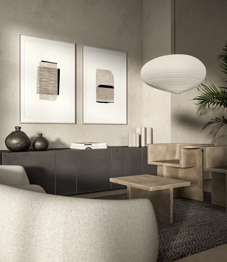 May include: Two framed abstract art prints with beige and black paper textures hang on a light beige wall above a dark gray cabinet. A white, round, layered pendant light hangs from the ceiling. A white, curved sofa sits in front of the cabinet. A light brown, rectangular coffee table sits in front of the sofa. A light brown, abstract sculpture sits on the floor.