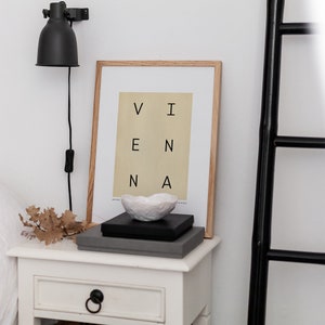 May include: A white nightstand with a drawer and a black knob. On top of the nightstand is a framed print with the word "VIENNA" in black letters on a light yellow background. There is a small white ceramic bowl on top of a black and gray box.