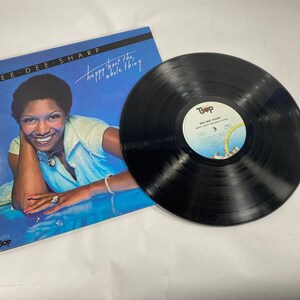 May include: A vinyl record album by Dee Dee Sharp, titled "happy about the whole thing." The album cover features a smiling woman in a blue outfit against a blue background. The record itself is black with a white and rainbow label.
