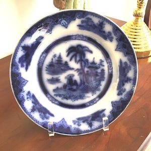 May include: A blue and white porcelain plate with a decorative design of a palm tree and other foliage. The plate has a scalloped edge and is displayed on a wooden surface.