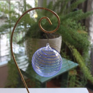 May include: A clear glass sphere with blue spiral stripes hangs from a decorative gold-colored metal stand. The sphere is suspended in front of a blurred background of greenery and a window, creating a delicate and artistic display.