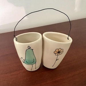 May include: Two off-white ceramic vases with a black wire handle. One vase features a teal bird illustration, the other a yellow flower. The vases are suitable for small plants or decorative use.