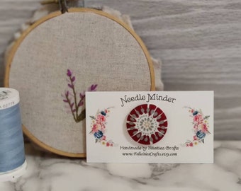 May include: A red and white needle minder with a floral design. The needle minder is handmade by Felicities Crafts and is available on Etsy.