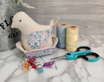 May include: A beige bird-shaped pincushion with a floral fabric design sits on a white marble surface.  Two spools of thread, one blue and one yellow, are next to the pincushion.  A pair of turquoise scissors and a variety of colorful sewing clips are also on the surface.