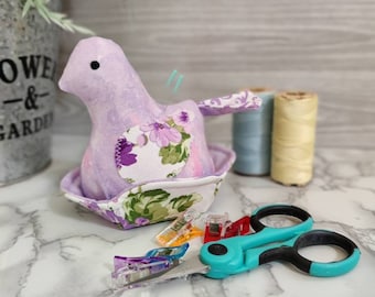 May include: A purple and floral fabric pincushion shaped like a bird sitting in a nest. The pincushion is surrounded by sewing supplies, including scissors, thread spools, and clips.