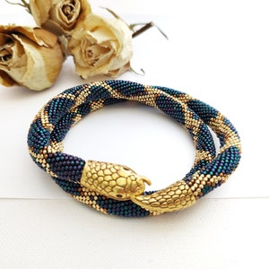 May include: A flexible beaded bracelet featuring a snake design with iridescent dark blue and purple scales alternating with metallic gold sections. The bracelet ends in a detailed gold-toned metal snake head and tail clasp that meet.