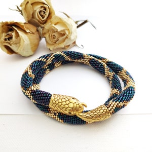 May include: A beaded bracelet in a snake design. The bracelet is primarily dark blue and gold, with a snake head clasp. The bracelet is coiled and lies on a white surface, with dried flowers in the background.