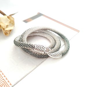 May include: A flexible, tubular mesh bracelet featuring a silver-toned snake head clasp. The bracelet displays a gradient colour transition from a light, metallic silver at one end to a dark, charcoal grey at the other, coiled into a circular shape.
