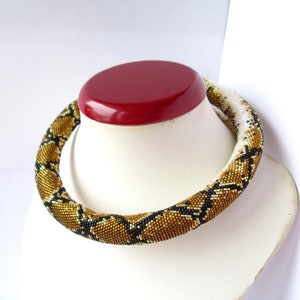 May include: A tubular beaded necklace featuring a repeating geometric pattern in shades of metallic gold, black, and white. The necklace is constructed from small, uniform glass beads arranged in a dense, cylindrical structure that encircles the neck.