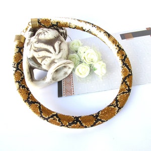 May include: A tubular beaded necklace featuring a repeating snake-skin pattern in shades of metallic gold, black, and white. The necklace is finished with ribbed bronze-toned metal end caps and a small lobster claw clasp at the back.