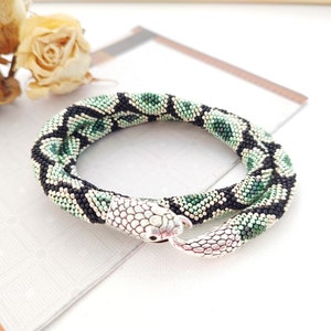 Puede incluir: Un collar de serpiente de cuentas verdes y negras con un cierre de cabeza de serpiente plateado.