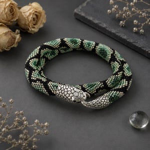May include: A flexible beaded bracelet featuring a snake-like design with a silver-toned metal head and tail clasp. The body is crafted from small beads in a repeating pattern of black, forest green, and light green, mimicking a serpent's scales.