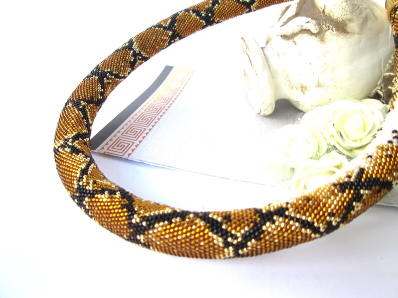 May include: A tubular necklace crafted from tiny glass beads in a repeating diamond pattern. The beads are colored in metallic bronze, gold, and black, creating a textured surface that mimics a snake skin design. The necklace is flexible and rounded.