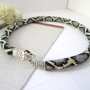 May include: A circular beaded necklace featuring a repeating diamond pattern in black, grey, and metallic gold. The necklace ends in a silver-toned metal snake head clasp that connects to the tail, creating a continuous serpent-like design.
