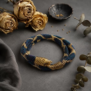 Puede incluir: Una pulsera flexible con forma de serpiente hecha de cuentas de semillas azul oscuro iridiscente y doradas. La pieza presenta un cierre de metal dorado detallado con cabeza y cola que se unen para formar un bucle circular continuo.