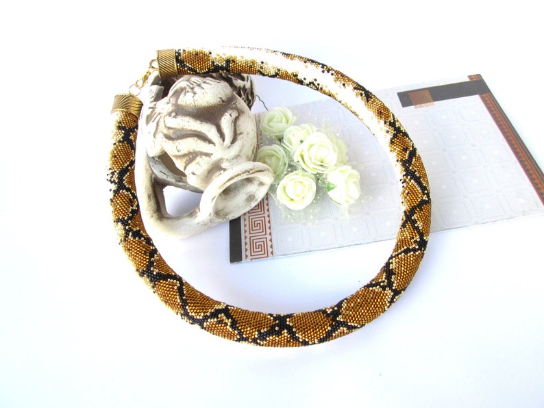 May include: A tubular beaded necklace featuring a repeating snake-skin pattern in shades of metallic gold, black, and white. The necklace is finished with ribbed bronze-toned metal end caps and a small lobster claw clasp at the back.