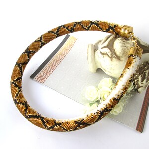 May include: A tubular beaded necklace featuring a repeating diamond pattern in shades of metallic bronze, black, and white. The necklace is finished with gold-toned metal end caps and a lobster claw clasp, creating a flexible, rounded structure.