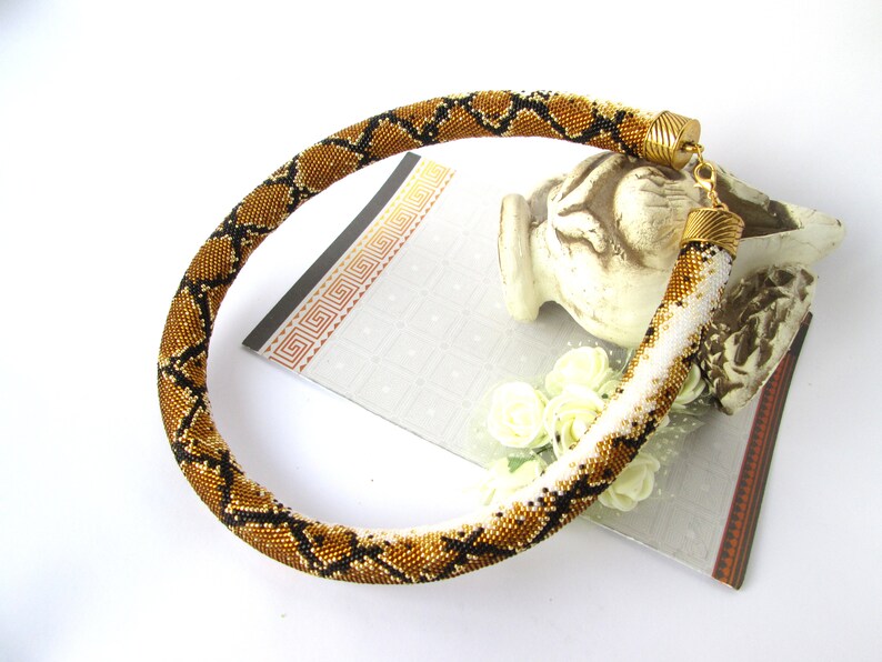 May include: A tubular beaded necklace featuring a repeating diamond pattern in shades of metallic bronze, black, and white. The necklace is finished with gold-toned metal end caps and a lobster claw clasp, creating a flexible, rounded structure.