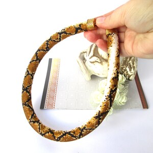 May include: A tubular beaded necklace featuring a repeating snake-skin pattern in shades of metallic gold, black, and white. The necklace has a cylindrical shape with a textured surface made of tiny glass beads and is finished with gold-toned metal end caps.