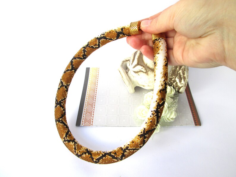 May include: A tubular beaded necklace featuring a repeating snake-skin pattern in shades of metallic gold, black, and white. The necklace has a cylindrical shape with a textured surface made of tiny glass beads and is finished with gold-toned metal end caps.