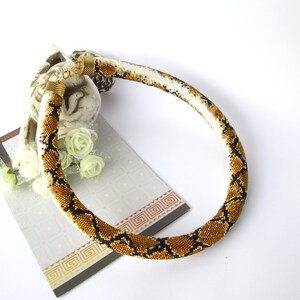 May include: A tubular beaded necklace featuring a repeating snake skin pattern in shades of gold, black, and white. The necklace is finished with metallic gold-toned end caps and a lobster claw clasp, creating a continuous circular shape.