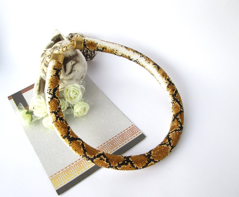 May include: A tubular beaded necklace featuring a repeating snake skin pattern in shades of gold, black, and white. The necklace is finished with metallic gold-toned end caps and a lobster claw clasp, creating a continuous circular shape.