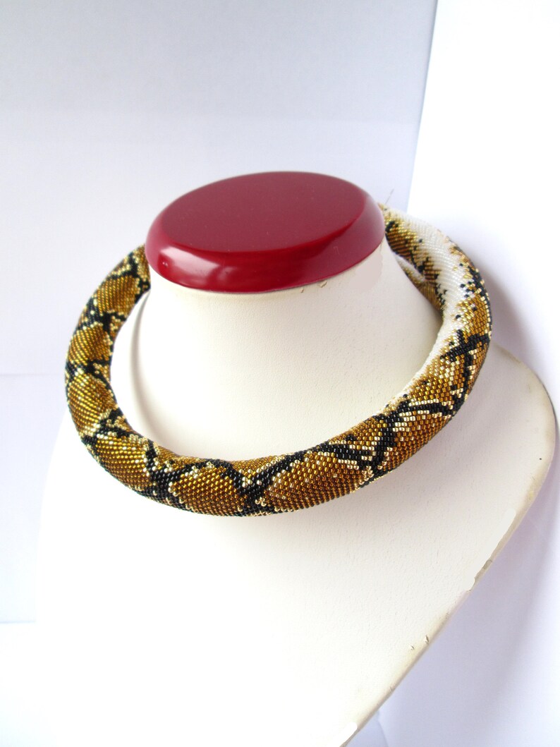 May include: A tubular beaded necklace featuring a repeating geometric pattern in shades of metallic gold, black, and white. The necklace is constructed from small, uniform glass beads arranged in a dense, cylindrical structure that encircles the neck.