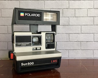 May include: A silver Polaroid Sun 600 instant camera with a black top and a red button. The camera has a built-in flash and a light management system.