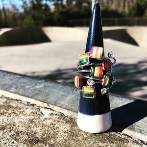 May include: A stack of colorful rings with wood inlay and silver settings on a white and blue display stand. The rings are stacked on top of each other and have a variety of colors and patterns.