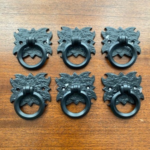May include: Six black metal drawer pulls with ornate designs. Each pull has a ring-shaped handle.