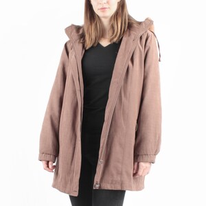 Könnte beinhalten: Eine Frau trägt eine braune Kapuzenjacke über einem schwarzen Oberteil und schwarzen Jeans. Die Jacke hat einen Knopfverschluss und die Frau trägt braune Lederstiefel. Der Hintergrund ist weiß.