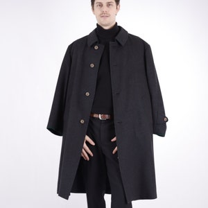 Könnte beinhalten: Ein dunkelgrauer Mantel mit Knopfleiste, getragen über einem schwarzen Rollkragenpullover und einer schwarzen Hose. Der Mantel hat ein klassisches Design mit Kragen und braunen Knöpfen. Das Outfit wird durch einen braunen Gürtel und braune Schuhe ergänzt.