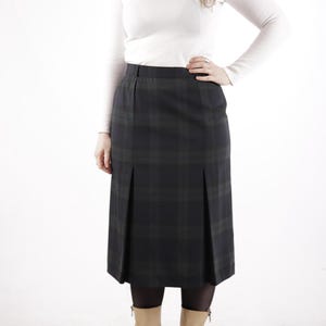 May include: A black and green plaid skirt with a high waist and a straight cut. The skirt has pleats in the front and a zip at the back. The skirt is knee-length and is being worn with a white long-sleeved shirt and tan boots.