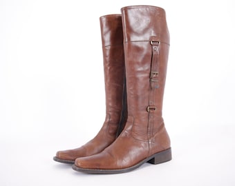 May include: A pair of brown leather knee-high boots with buckle accents and a zipper on the inside of the boot.