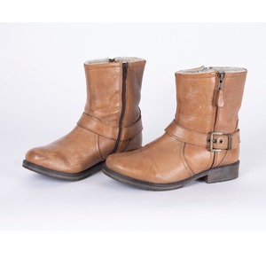 Könnte beinhalten: Ein Paar braune Leder-Ankle Boots mit seitlichem Reißverschluss und Schnallen Detail. Die Stiefel haben ein Futter aus Kunstfell und einen niedrigen Absatz. Die Stiefel werden auf weißem Hintergrund präsentiert.