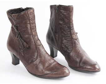 May include: A pair of brown leather ankle boots with a small tassel detail on one boot. The boots have a low heel and a pointed toe.