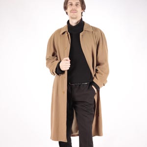 May include: A long, brown wool coat with a button closure and a collar. The coat is worn over a black turtleneck sweater and black pants.
