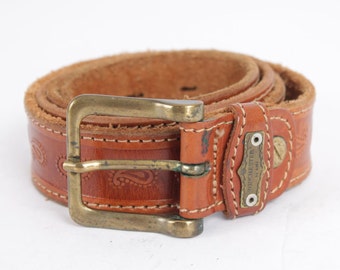 May include: A brown leather belt with a paisley pattern and a brass buckle. The belt has a label that reads "WINCHESTER by AMI".