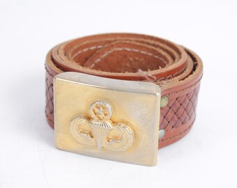 May include: A brown leather belt with a silver and gold buckle featuring a parachute design. The belt has a woven texture on the edges and is rolled up. The buckle is rectangular.