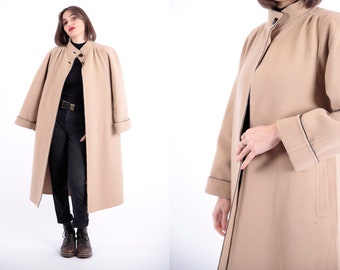 May include: A long, beige wool coat with a black trim. The coat has a high collar and a button closure. The coat is shown on a model wearing black pants and boots.
