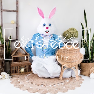 May include: A person in a white bunny costume with a blue jacket and a bow tie sits near a wicker chair and a decorative birdcage. The scene is set with plants, a woven basket, and a round jute rug. The text "eclectic existence photography" is visible.